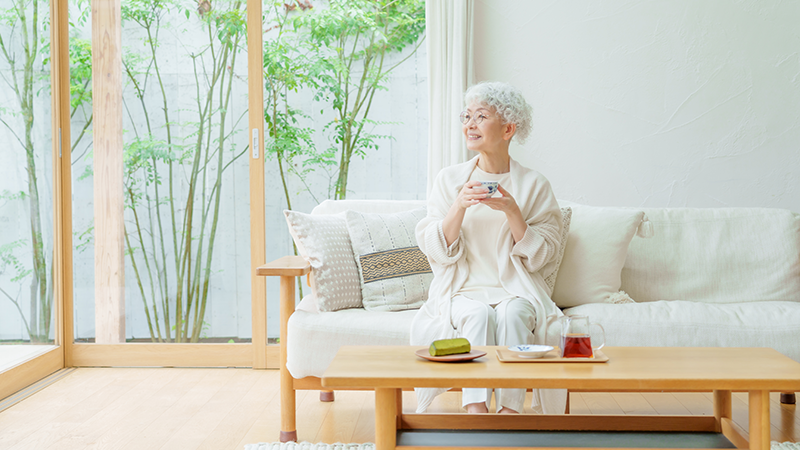自宅のソファーでリラックスしてお茶を飲む、笑顔の日本人高齢女性のイメージ